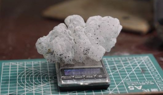 A spiky white hailstone, flecked with small dots of black paint, rests on a scale on a desk. 